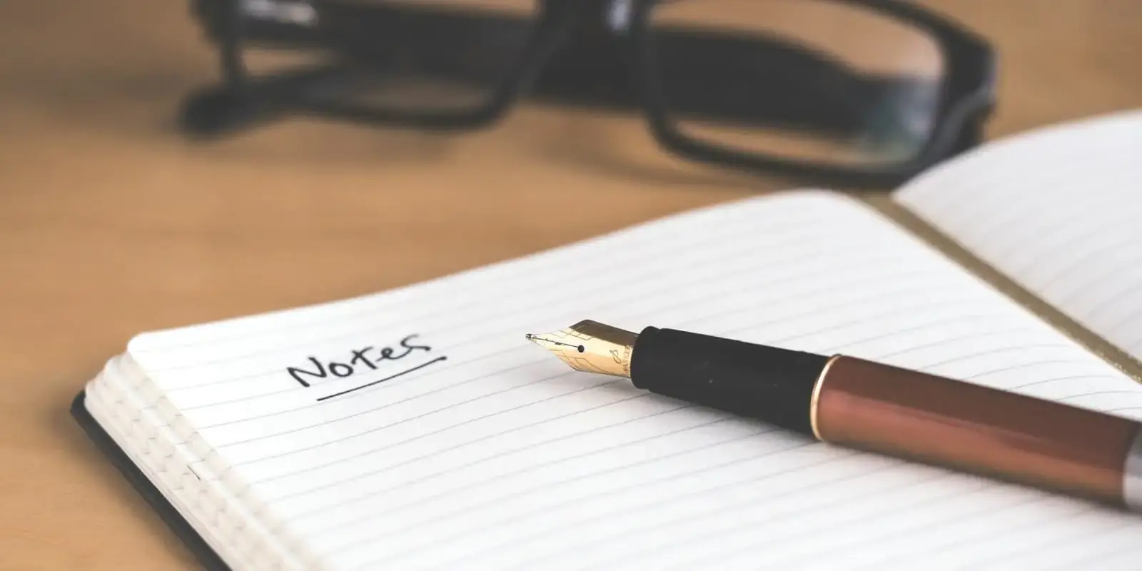 Cuaderno abierto con la palabra Notes escrita, una pluma y unas gafas sobre una mesa